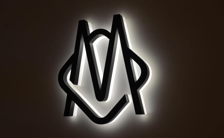 Black monogram logo in 3D casting a white halo onto a dark wall